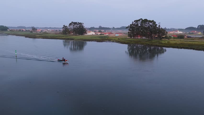 Aerial view of a small boat navigating the calm waters of Aveiro Lagoon with lush green banks and trees under an overcast sky in Aveiro District, Portugal.