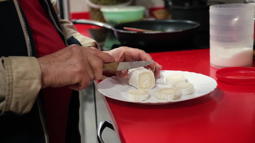 Senior male hands slicing a fresh log of goat cheese on a white plate in a modern kitchen