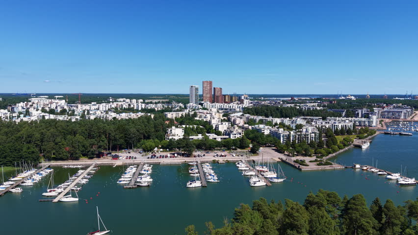Aerial view of Vuosaari marina in Helsinki, Finland, featuring sailboats moored at docks, modern residential towers, green forest and a second marina along the calm waterfront