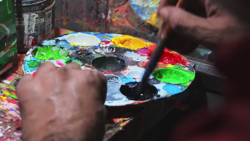 Close-up view shows a Dhaka artist using a paintbrush to mix bright acrylic pigments on a colorful palette wheel, preparing paints to document an endangered rickshaw art custom in Bangladesh.