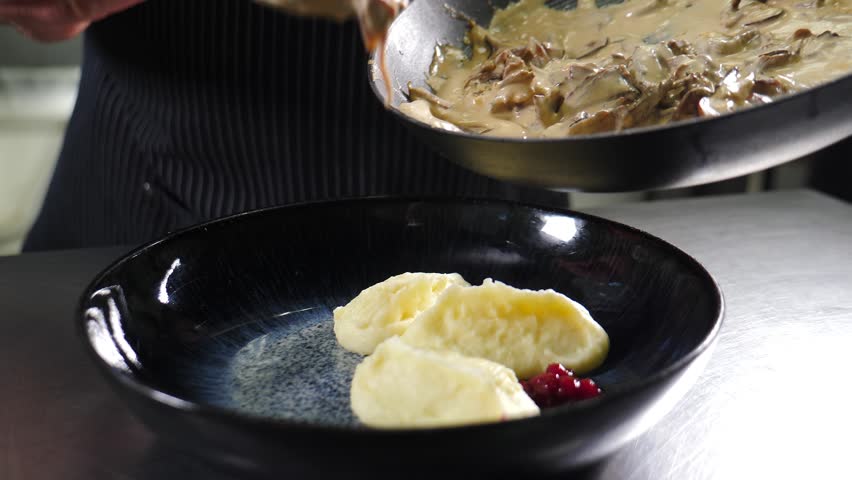 Chef hand pouring thick savory meat sauce over gourmet dish served with potatoes and red cranberry garnish on black plate. Close-up fine dining plating. 4k video