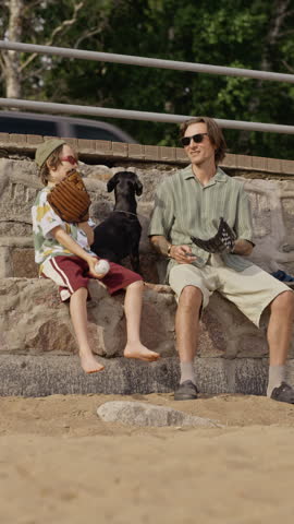 Vertical video of Caucasian father and young son sitting on stone steps by beach with dog, playing with baseball. Footage is perfect for promoting family bonding and outdoor summer activities