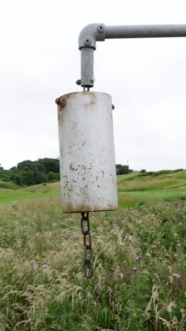 Old, rusty white bell swinging in the wind over a field of tall grass on an overcast day