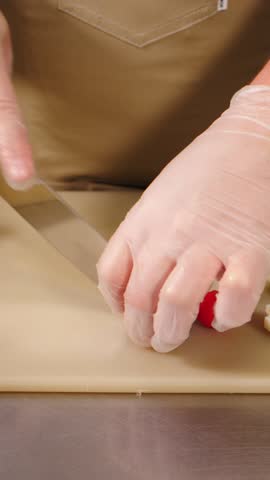 Close-up of chef gloved hands carefully slicing fresh red cherry tomato with small knife on cutting board, surrounded by boiled egg halves. Food prep. Restaurant food cooking. vertical 4k video
