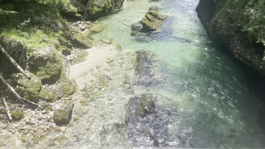 Aerial-style POV view from a bridge overlooking a wild river as a group of rafters paddles through whitewater rapids. Eight people wearing helmets navigate between moss-covered rocks surrounded by natural vegetation. Adventure sports and outdoor recreation concept.