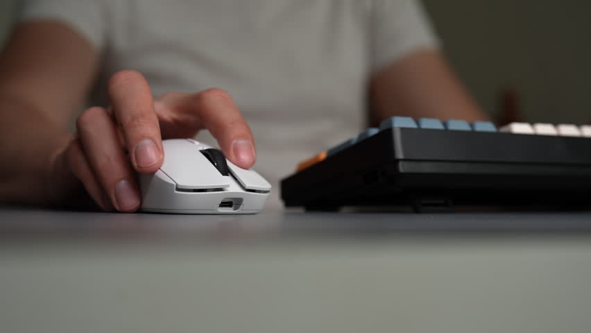 Close-up of person hand using white wireless mouse. Fingers click and scroll. Another hand appears to type on mechanical keyboard. Modern office, gaming setup for work, play. Shooting in slow motion.