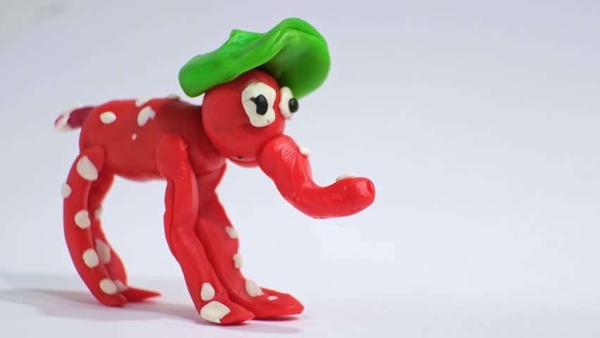 A small red plasticine elephant with white spots and a green hat stands in a studio on white background as a strawberry. Imagination and fine motor skills. Handmade children