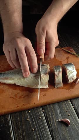 A person cuts fish on a wooden board in a home kitchen. The setting shows hands skillfully slicing the fish into pieces. The kitchen has a simple design with dark surfaces.