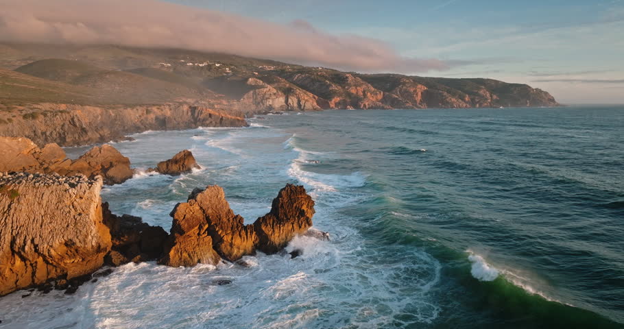 Rugged coastline of Sintra Cascais Natural Park with dramatic ocean waves crashing against rocks and towering cliffs with golden light during a beautiful sunset. Travel Portugal. Aerial drone panorama