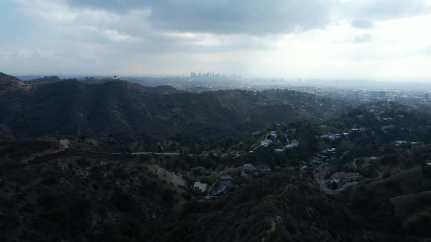 A moody aerial landscape of rolling hills with a distant city skyline beneath a cloudy sky