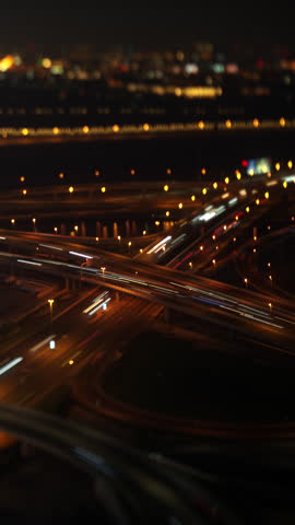Highway interchange in Dubai at night, car light trails curve across the stacked flyovers, street lamps line the ramps, water and city lights sit behind, tilt shift miniature look, timelapse