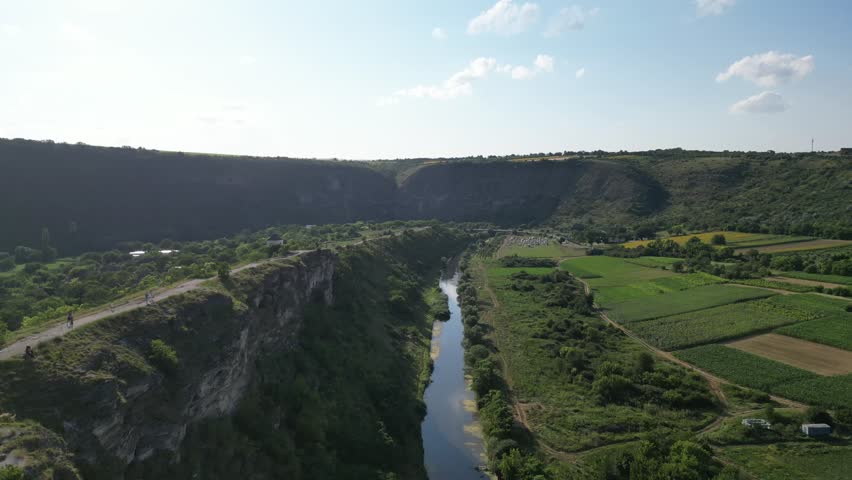 Aerial perspective of a deep river valley with steep cliffs. The river flows through green fields and hills. Farm land stretches along the riverbank. Clouds dot the sky