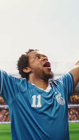 Vertical frame: African American football player in blue jersey cheering and saluting fans, entering stadium pitch before game, with determined footballer showing team spirit.