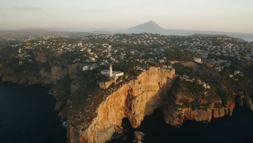 A drone view of the Alicante cliffs, Spain at sunrise, featuring a lighthouse perched above rugged rock formations and distant mountain scenery