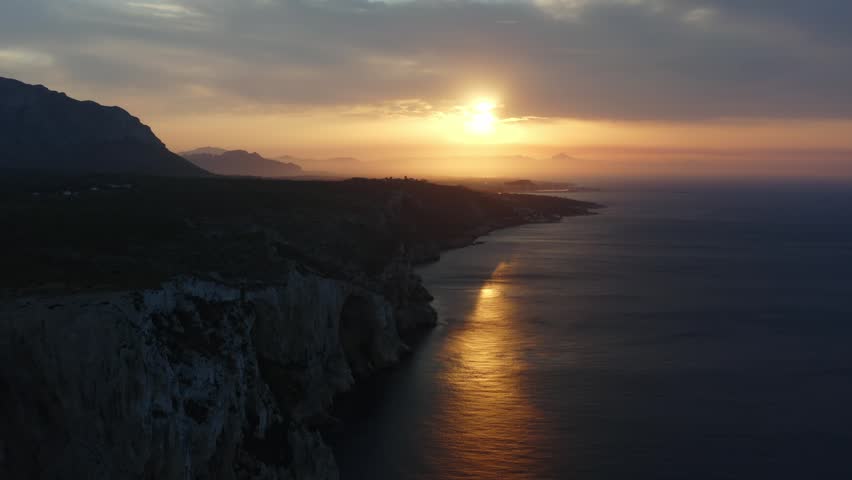 A drone view of the Alicante coastline, Spain at sunrise, with rugged cliffs, glowing reflections on calm water, and misty mountains