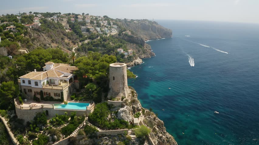 An aerial view of Javea, Xabia, Alicante, Spain, showing a historic watchtower, luxury cliffside villas, and turquoise Mediterranean waters below