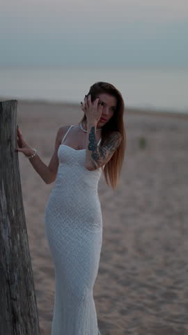 Elegant woman in white lace dress posing on sandy beach beside weathered wooden post at dusk