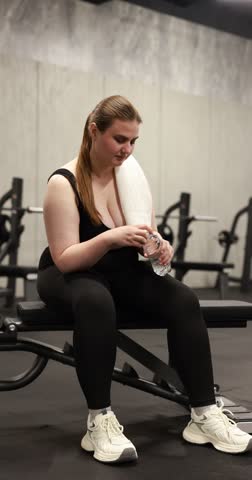 Overweight woman with bottle of water sitting on equipment in gym