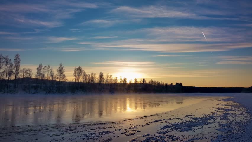 A vibrant sunset over a half-frozen misty lake reflects orange light, lined by frosted bare trees.