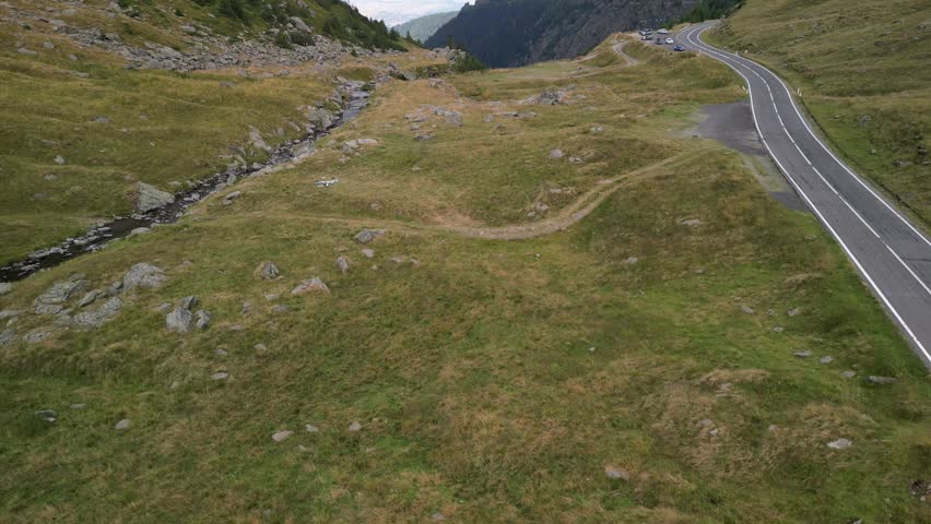 A drone view of mountain stream runs through a grassy, rocky valley next to a winding road and parking area.