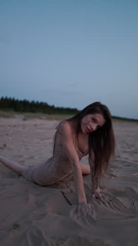 Young woman kneeling on sandy beach at dusk with long hair, fitted knit dress, and tattooed arm under calm twilight sky