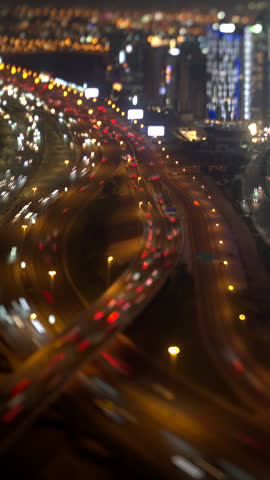 Dubai at night, red tail light trails pack the highway lanes, camera pans left to right over the lit towers, tilt shift lens gives a miniature look, vertical timelapse