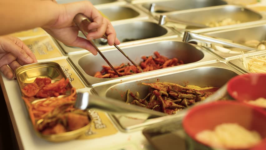 Close-up of hands selecting various fermented side dishes from metal trays in a restaurant.