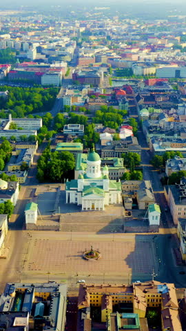 Vertical video. Helsinki, Finland. Helsinki Cathedral dominates the northern edge of Senate Square from an aerial perspective, its white stucco exterior and distinctive green copper dome rising above the neoclassical ensemble. The cathedral was designed by Carl Ludvig Engel in the Empire style, a variant of neoclassical architecture, and was completed in 1852 after more than two decades of construction. Its Greek cross plan features six Corinthian columns on each of the four identical facades, while twelve zinc statues of the Apostles adorn the roofline pediments. The surrounding Senate Square presents a cohesive neoclassical urban composition, with the Government Palace and University of Helsinki main building flanking the open plaza. The scene is captured in bright daylight under clear summer skies, with the Baltic Sea visible beyond the dense historic center and modern harbor infrastructure in the distance. The aerial camera movement reveals the geometric order of the grid-planned capital, the contrast between the cathedral
