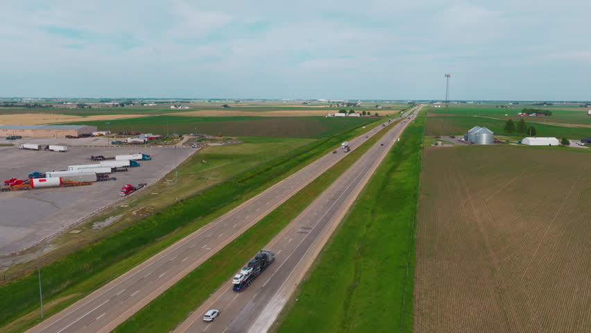 Commercial car hauler truck transporting vehicles down an interstate highway in rural Iowa next to a major truck stop logistics facility.