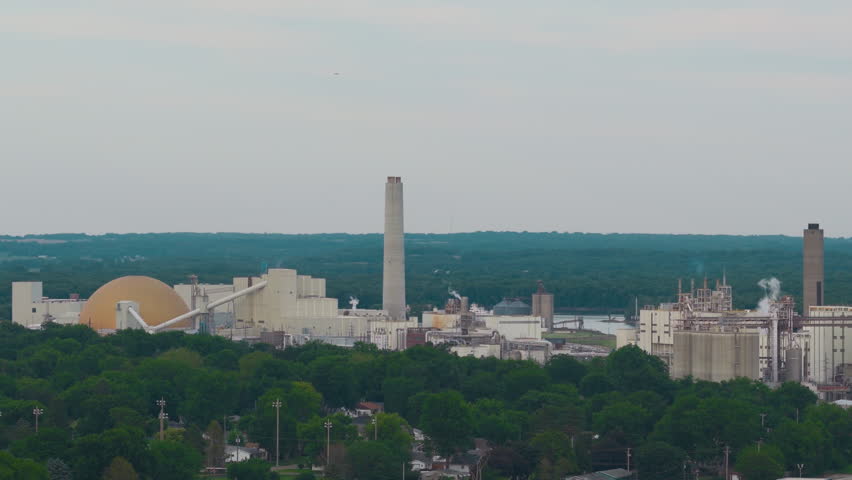 Aerial of Clinton, Iowa: a large grain processing complex with golden dome, silos and smokestack rises over tree-lined neighborhoods near the Mississippi River.