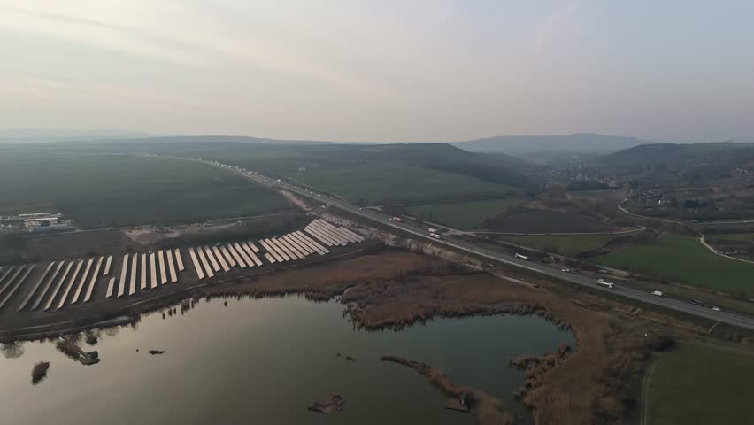 Bicske, Hungary motorway traffic with cars and trucks passing solar panels, farmland and a fishing lake in the rural landscape, captured in an aerial view.