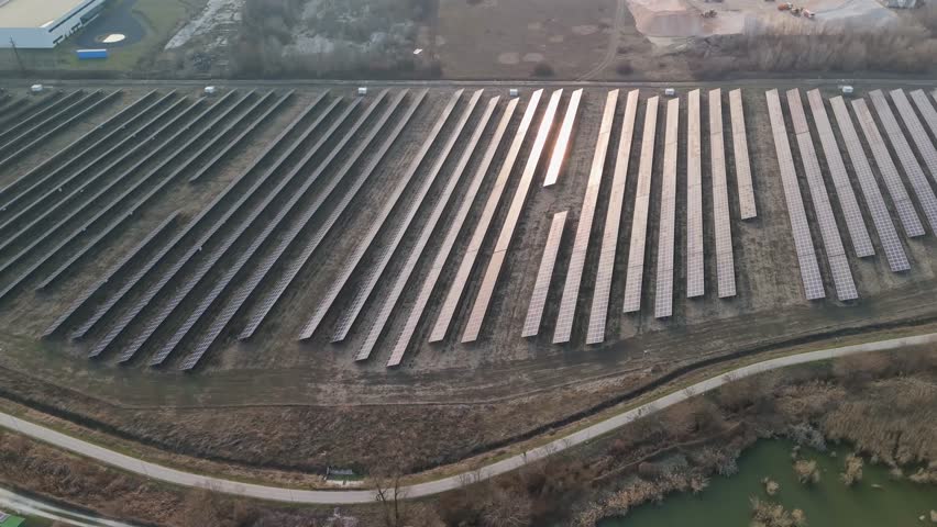 Bicske, Fejér County, Hungary solar farm with photovoltaic arrays beside fields, trees and a pond at sunset, utility-scale renewable energy infrastructure tracked by drone.