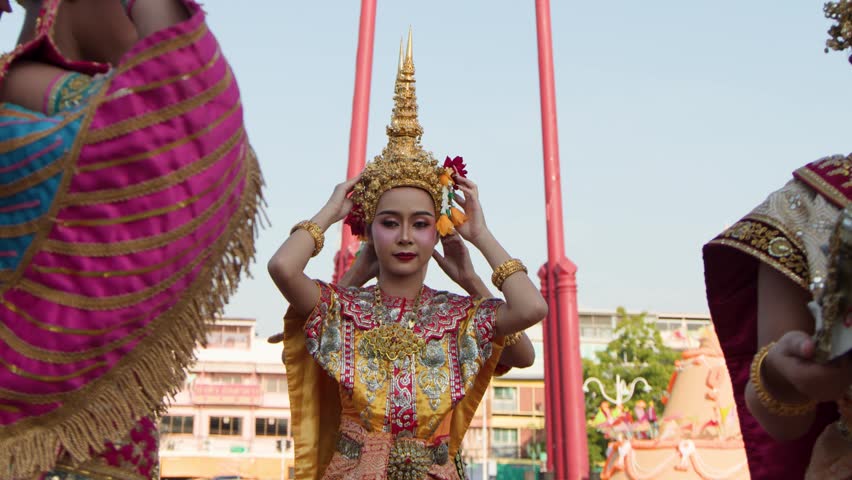 Female performers in ornate traditional costumes adjust golden headdresses during a bright outdoor cultural event.