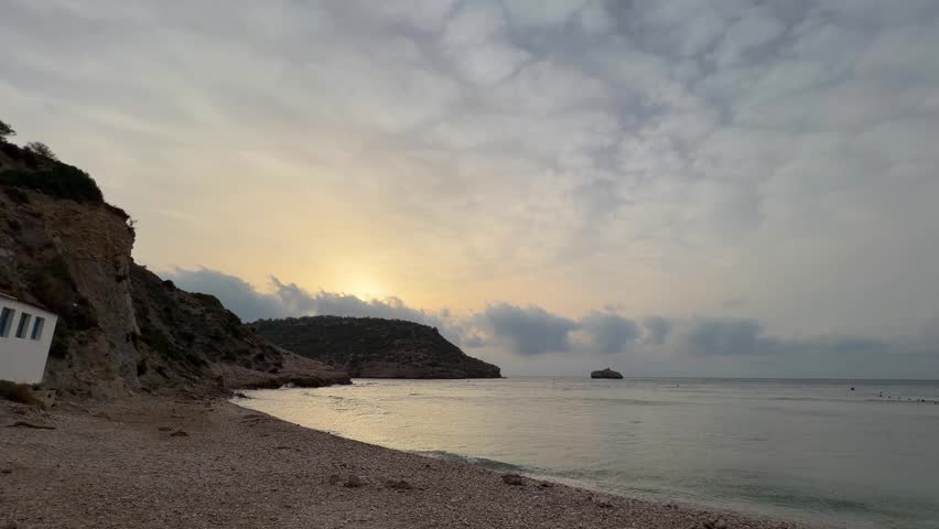 Sunrise on Rocky coastline of Cala Portixol in Jávea, Spain, featuring traditional white Mediterranean buildings and a pebble beach under a dramatic cloudy sky at sunrise, reflecting on the tranquil s