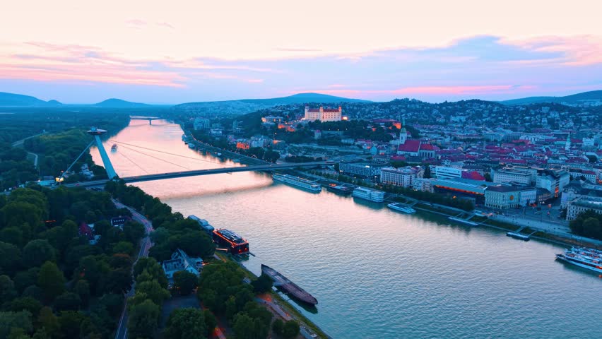 High aerial view of Bratislava cityscape, Danube river, and castle at twilight. Historic buildings, cable bridge, and waterfront illuminated by evening light in Slovakia.