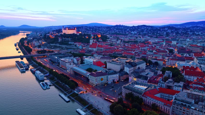 Aerial view of Bratislava old town waterfront and castle at dusk. Riverbank streets, historical buildings, and hill fortress lit up during twilight in Slovakia.