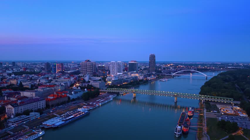Aerial view of Danube river and Bratislava cityscape at dusk. Drone shot of illuminated bridges, modern skyscrapers and cruise ships during twilight.