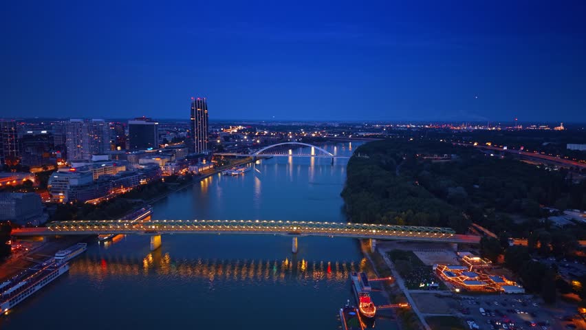 Night aerial skyline of Bratislava with bridges over Danube river. Illuminated Old Bridge and Apollo Bridge crossing water near tall modern buildings.