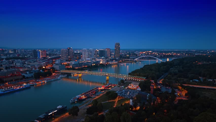 Wide aerial panorama of Bratislava city along Danube river. High perspective view of illuminated bridges, river banks and dark urban landscape, Slovakia.