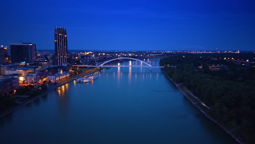 Night aerial view of illuminated arch bridge and river skyline. Night aerial view of illuminated arch bridge over river with dark blue sky, city lights, and forest in background.