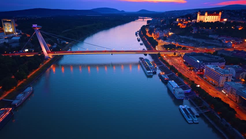 Aerial night view of illuminated Bratislava castle and cable bridge. Night aerial view of illuminated castle, cable stayed bridge over river, city lights, and docked ships in Bratislava.