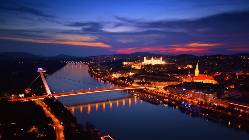 Panoramic view of Bratislava landmarks and Danube river at night. Wide drone perspective of illuminated bridge, castle and cathedral under dusk sky, Slovakia.