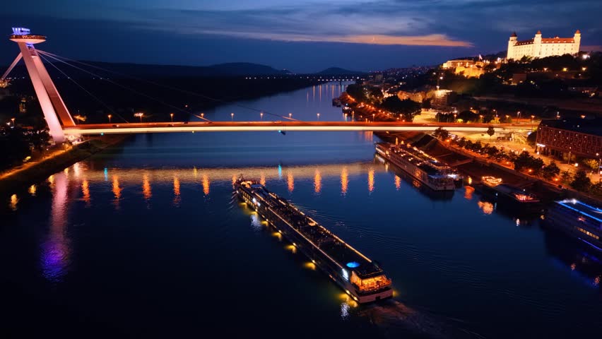 Aerial view of illuminated bridge and castle over river. Night aerial view of illuminated bridge, Danube river with passenger ship, and castle on hill in Bratislava.