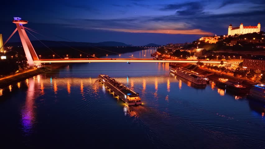 Night aerial view of Most SNP and Danube in Bratislava. Illuminated cruise boat moving on river near bridge tower with city reflections.