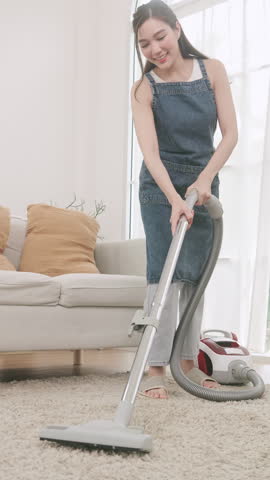 A woman using a canister vacuum cleaner to clean a fluffy carpet in a bright and cozy living room.