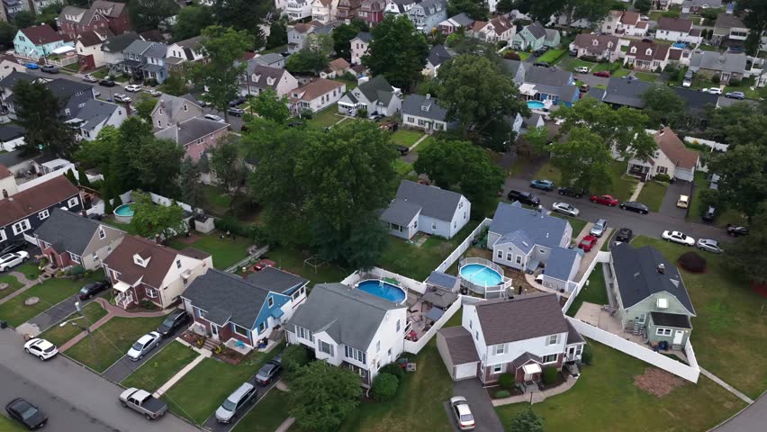 Round above-ground pools dot backyards of closely spaced houses in dense suburban neighborhood, mature trees plus parked cars lining streets in Belleville, New Jersey. Aerial drone top down