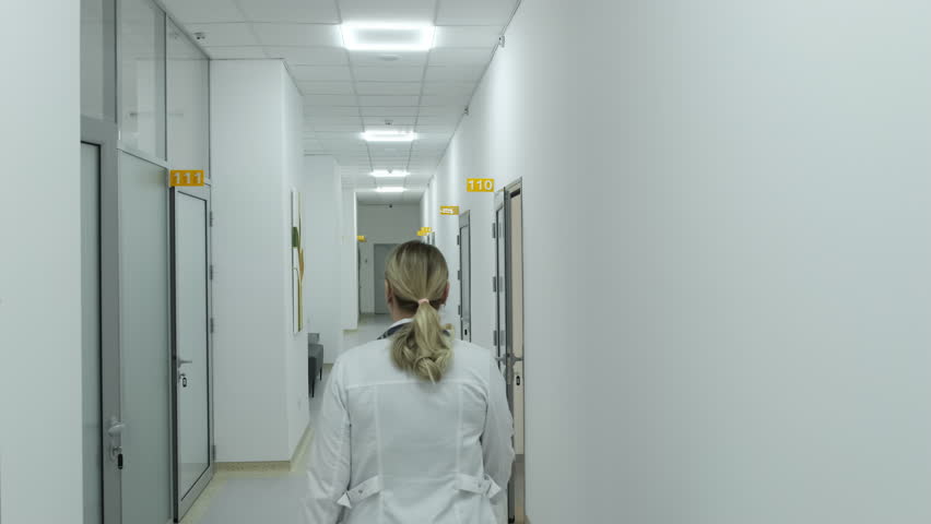 Doctor proceeds through contemporary corridor of healthcare facility. Woman prepares to provide medical care to patient. Slow motion