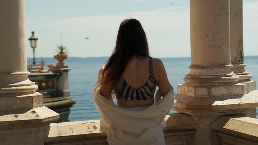 Young woman standing between stone columns overlooking the Mediterranean Sea from Miramare Castle in Trieste, Italy, on a bright summer day.