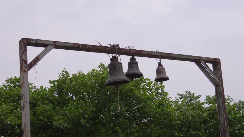 Three old bronze bells hanging on a rusty metal frame ringing loudly for a religious ceremony, with lush green trees and a cloudy sky in the background, signaling a call to worship