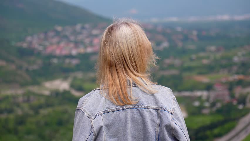 Blonde woman wearing a denim jacket standing and watching the scenic mountain valley, with her hair gently blowing in the wind. Beautiful green landscape with a small town in the background